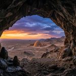صورة مولدة بالذكاء الاصطناعي تُظهر إطلالة من داخل كهف صخري طبيعي نحو بناء طيني تقليدي وسط صحراء واسعة وقت الغروب 