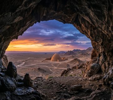 صورة مولدة بالذكاء الاصطناعي تُظهر إطلالة من داخل كهف صخري طبيعي نحو بناء طيني تقليدي وسط صحراء واسعة وقت الغروب 